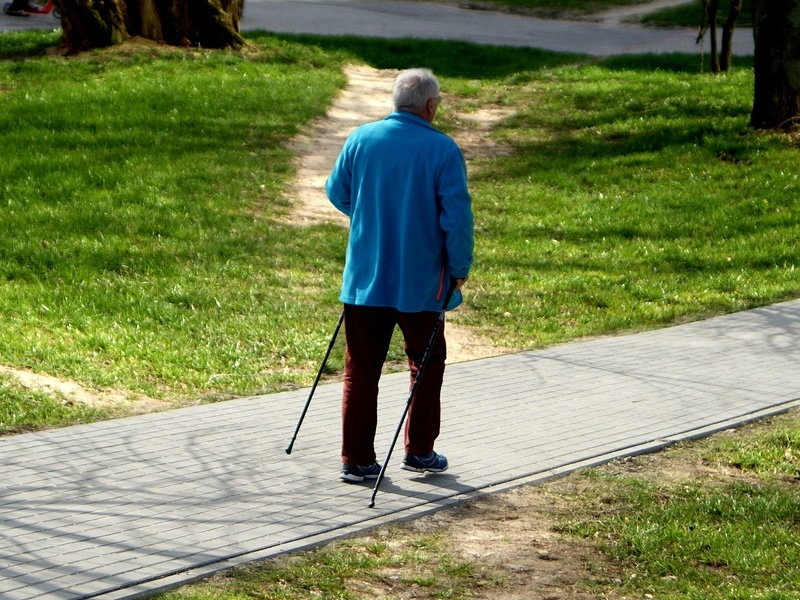 older adult walking on sidewalk with grass on both sides and trees in background