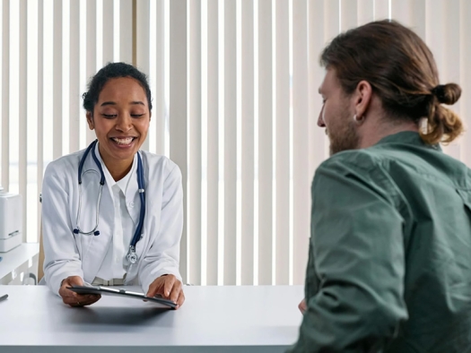 Female doctor and a young male patient sit across a desk from one another during a consultation in an office.