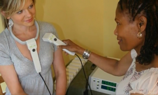 Two people seated at a small table. The person on the left wears part of the ONDAMED device around her neck and the person on the right holds a different part of the device on the person's chest near the heart.