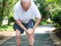 older man in white shirt, gray shorts, standing on paved road, in severe knee pain