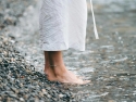 Person standing barefoot at edge of water on pebbly shore.