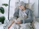 Woman in gray sweater and head scarf looking at phone while receiving an IV treatment sitting in bed with green plants on bedside table.