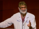Dr. Jamnadas, wearing white doctor's coat and black and gold surgical cap, standing a podium with microphone during his lecture