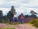 Two women walking on a wide dirt path towards trees in background 